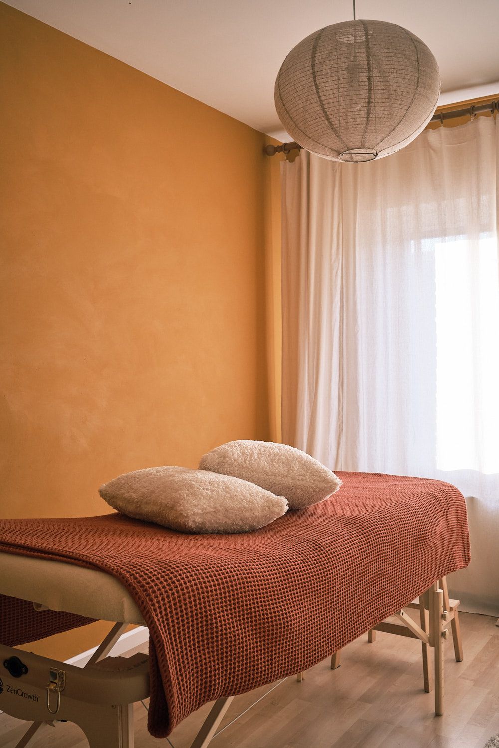 A view of the cocon jaune massage room, with a massage table in the middle, a lamp hanging above and a window behind it.