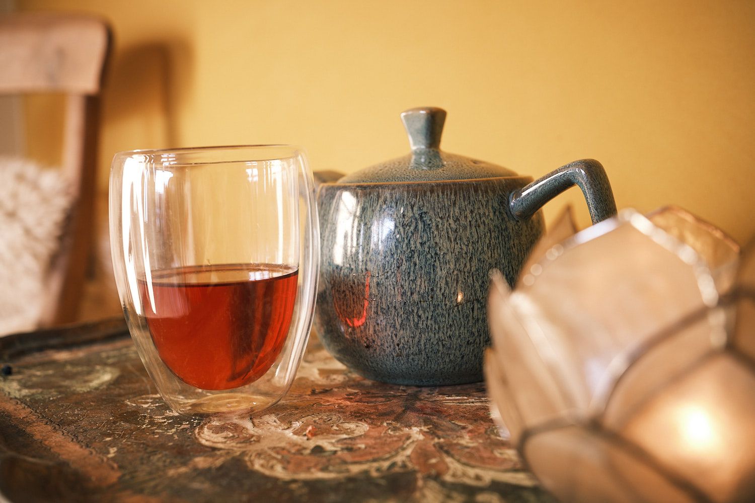 A glass mug with red tea and a blue teapot standing atop of a small table.